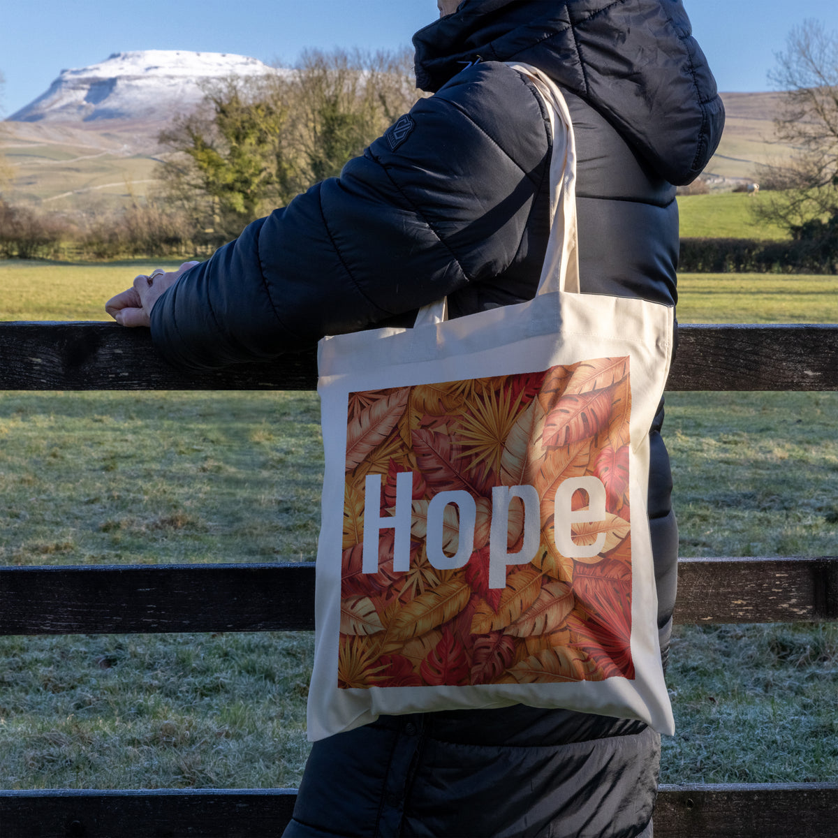 Person holding a tote bag with 'Hope' and autumn leaves design, standing in a field with a mountain in the background.
